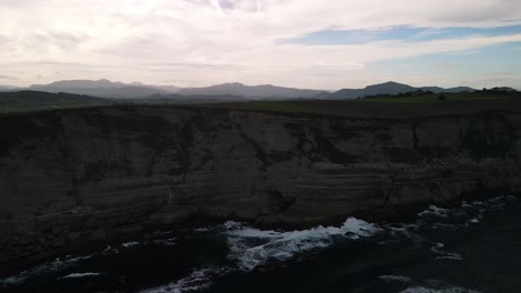 Hiker-walks-along-cliff-with-ocean-waves-crashing-on-rocks-at-shoreline