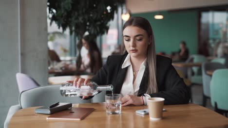 young,-attractive-woman-sitting-in-a-café,-dressed-in-business-attire,-pours-water-from-a-bottle-into-a-glass-and-drinks-it