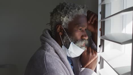 senior man with face mask on his chin looking out of window at home