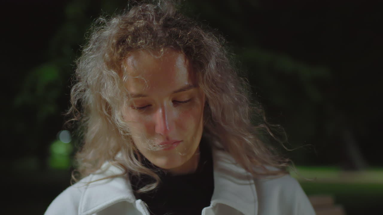 Lonely girl standing outdoors at night appearing deep in thought with eyes cast downward and wind gently swaying her curly hair, surrounded by soft green blur of night trees and calm atmosphere