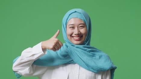 녹색 스크린 배경 스튜디오에 서 있는 아시아 무슬림 여성이 미소 짓고 엄지손가락을 올려 카메라에 제스처를 하고 있다.