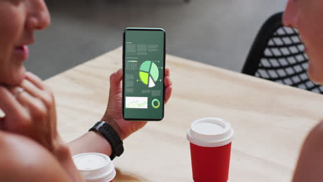 hombre y mujer caucásicos sentados a la mesa, usando un teléfono inteligente con estadísticas en la pantalla