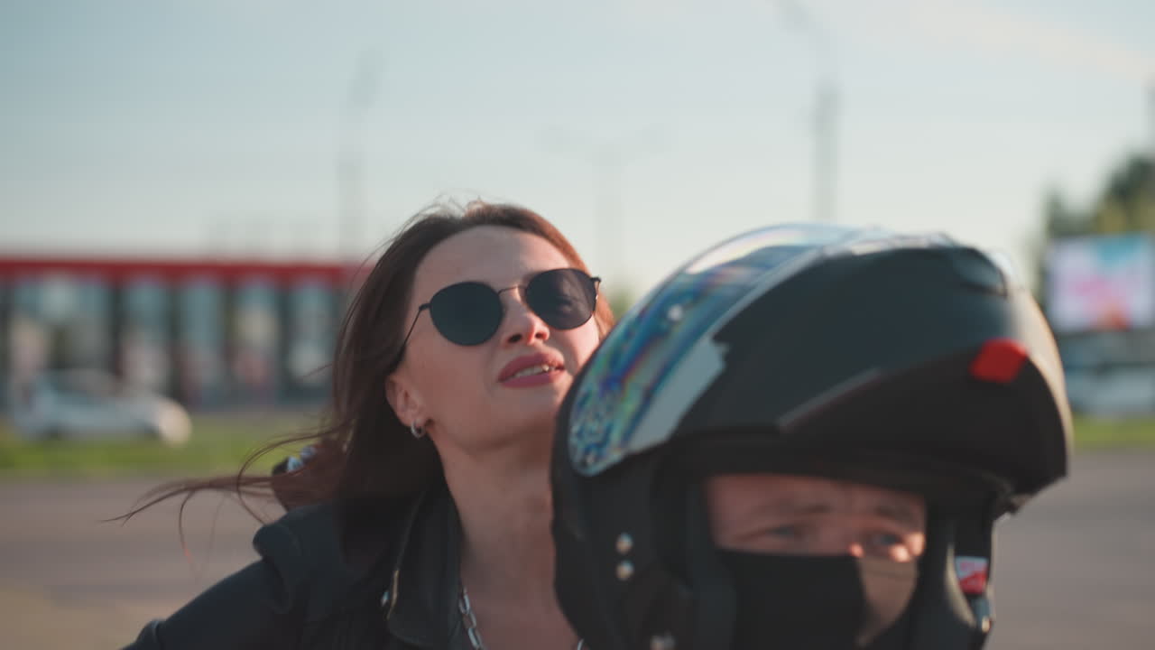 Close up of biker waiting for friend as woman holds helmet under arm climb behind, motorcycle mirror visible, blur background with trees and pavement