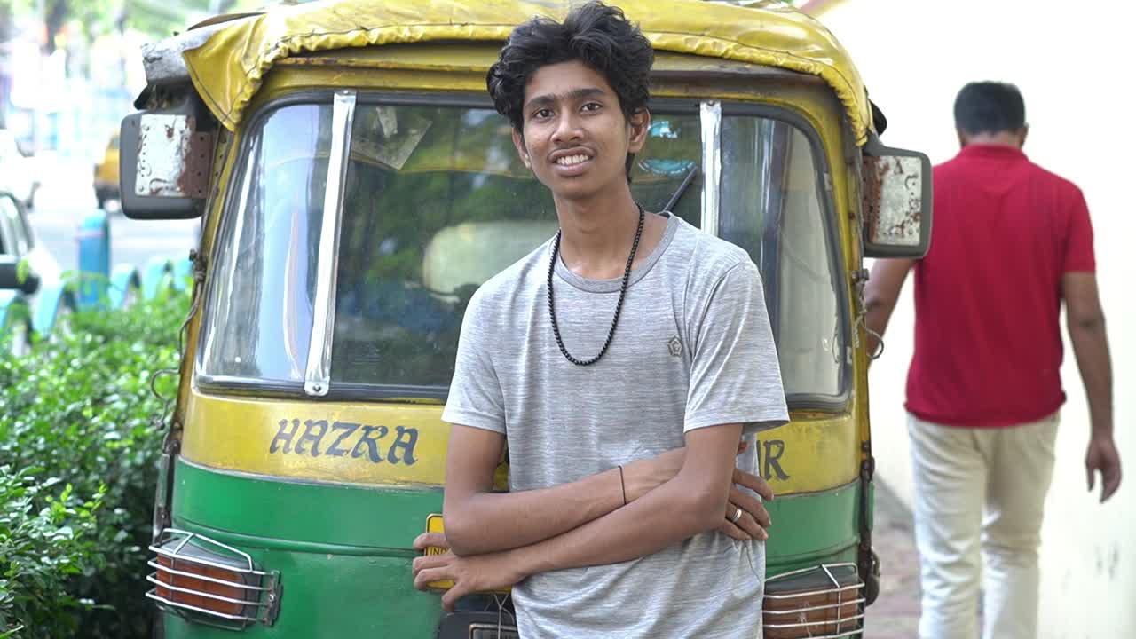 Slow motion shot of a young Indian auto driver in grey t-shirt standing in side of the road in front of the auto and giving a smile.