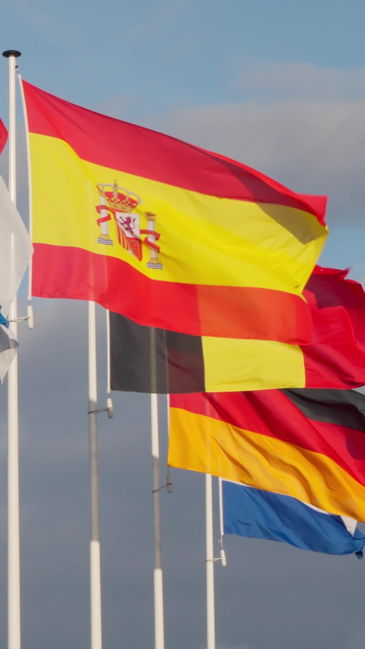 Several European national flags, including Spain, Belgium and Germany, wave gracefully against a cloudy blue sky. Flags represent various nations, showcasing international unity and diplomacy. Fabr...
