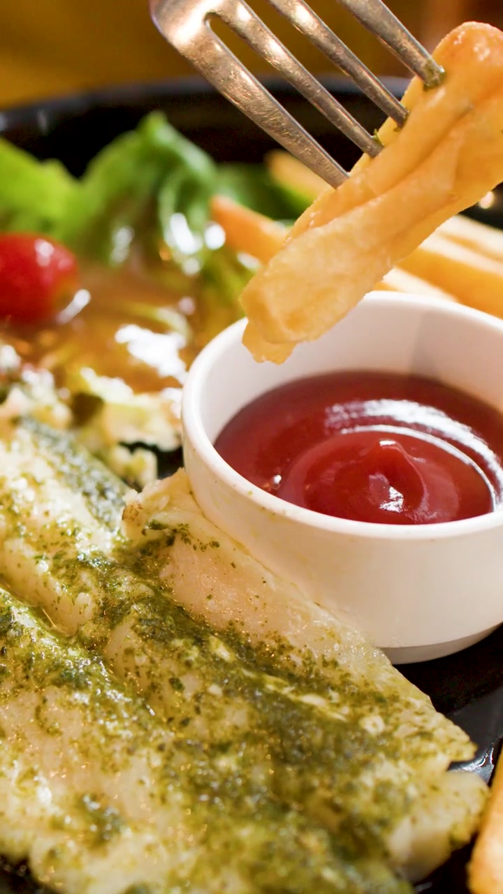 Un primo piano mostra una forchetta che intinge una patatina fritta nel ketchup accanto a pesce al pesto.
