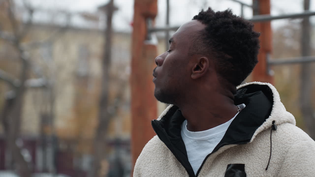 joven realizando ejercicios de torsión del cuello al aire libre durante el invierno, perdido en la concentración, el fondo muestra árboles desnudos, un edificio y un coche que pasa en el fondo borroso lejano