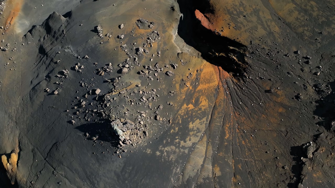 Top-down drone shot of dark volcanic ash and orange mineral streaks with scattered boulders, creating dramatic erosion patterns in Icelandic highland terrain.
