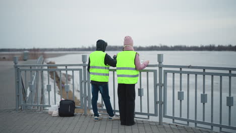 two people in bright reflective safety vests standing by riverside railing on cloudy day, observing calm water, with backpack and plastic bag on ground, peaceful scene of outdoor work or inspection pause