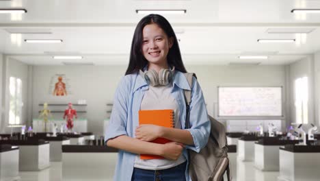 estudiante adolescente asiática con una mochila sosteniendo algunos libros y sonriendo a la cámara mientras está de pie en el laboratorio de ciencias