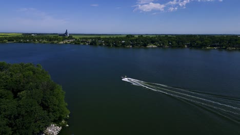 A-Recreational-Boat-Owner-Pulls-Two-Wakeboarding-People-Across-an-Undisturbed-Large-Natural-Freshwater-Lake-Against-a-Forest-Landscape-in-Killarney-Manitoba-Southern-Canada