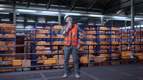cuerpo lleno de ingeniero masculino asiático con casco de seguridad de pie en el almacén con estantes llenos de productos de entrega. pensando en algo y mirando a su alrededor luego levantando su dedo índice