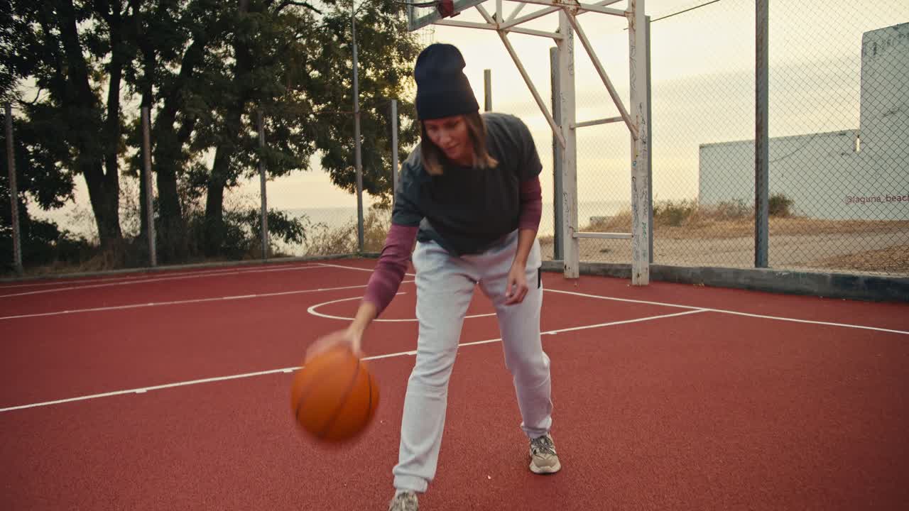 retrato de una chica con un peinado de bob en un sombrero negro que rebota una pelota de baloncesto naranja del suelo en una cancha de baloncestro y luego mira a la cámara y posa por la mañana durante su entrenamiento de balonceso