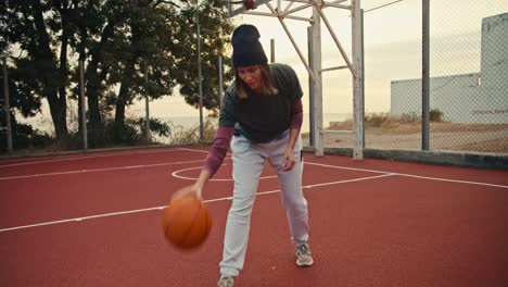 portrait of a girl with a bob hairstyle in a black hat who bounces an orange basketball off the floor on a basketball court and then looks at the camera and poses in the morning during her basketball training