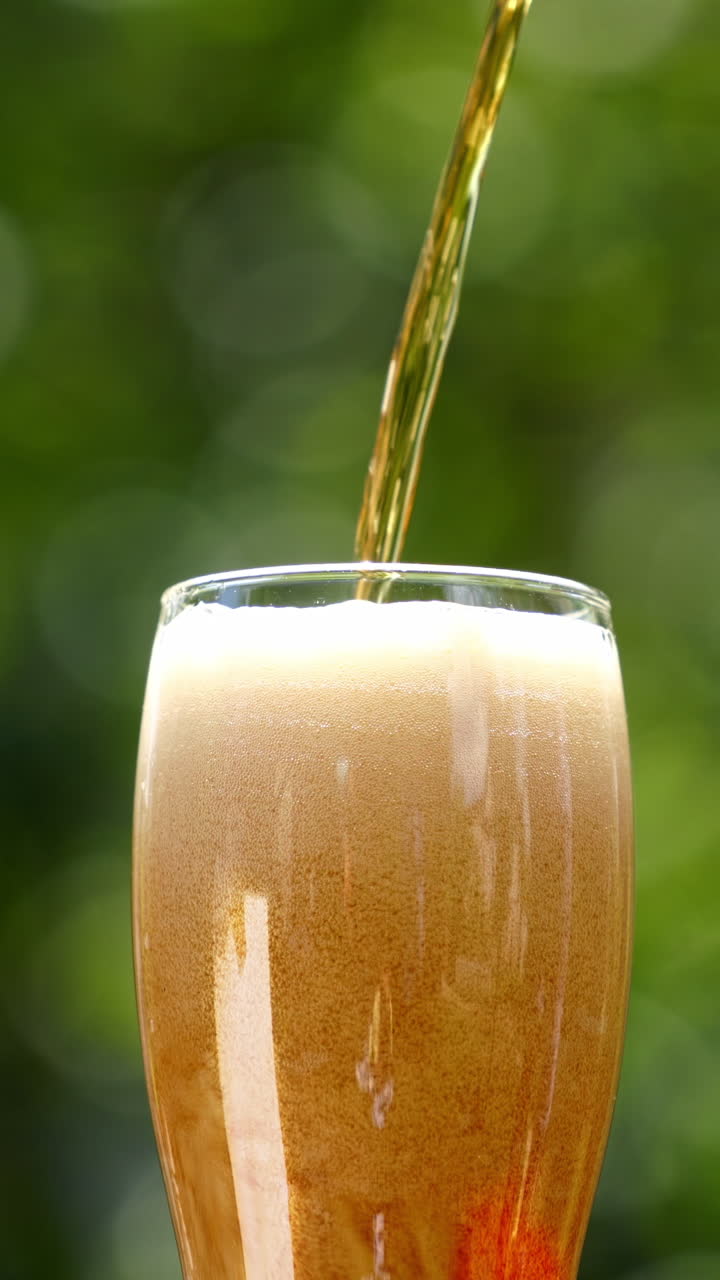 Pouring fresh beer with foam running down. Stream of beer pours into a beer glass against blur green background. Close-up. Vertical video