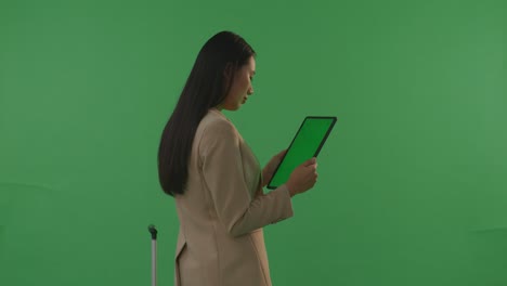 back view asian businesswoman traveler with luggage use green screen tablet while standing in the green screen background studio, during sunset time