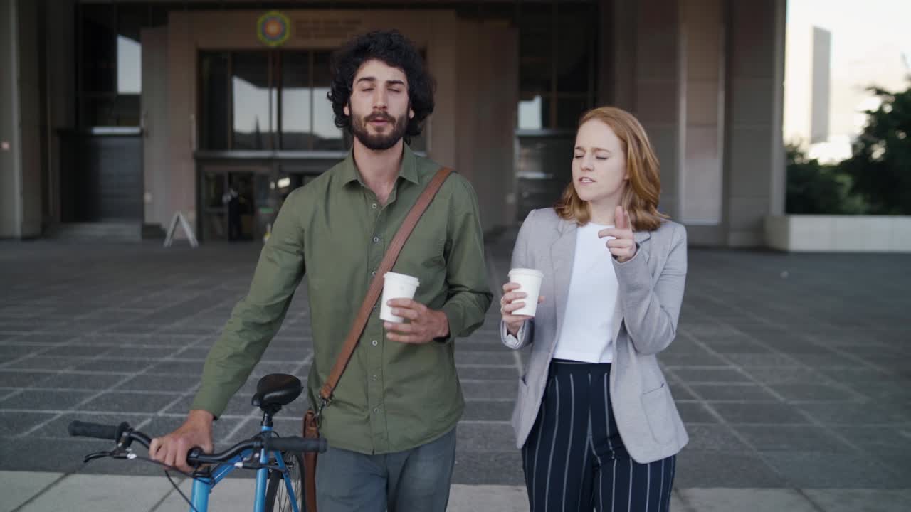 hombre caminando con su bicicleta y su amiga compañera de trabajo sosteniendo una taza de café frente al edificio corporativo hablando y sonriendo