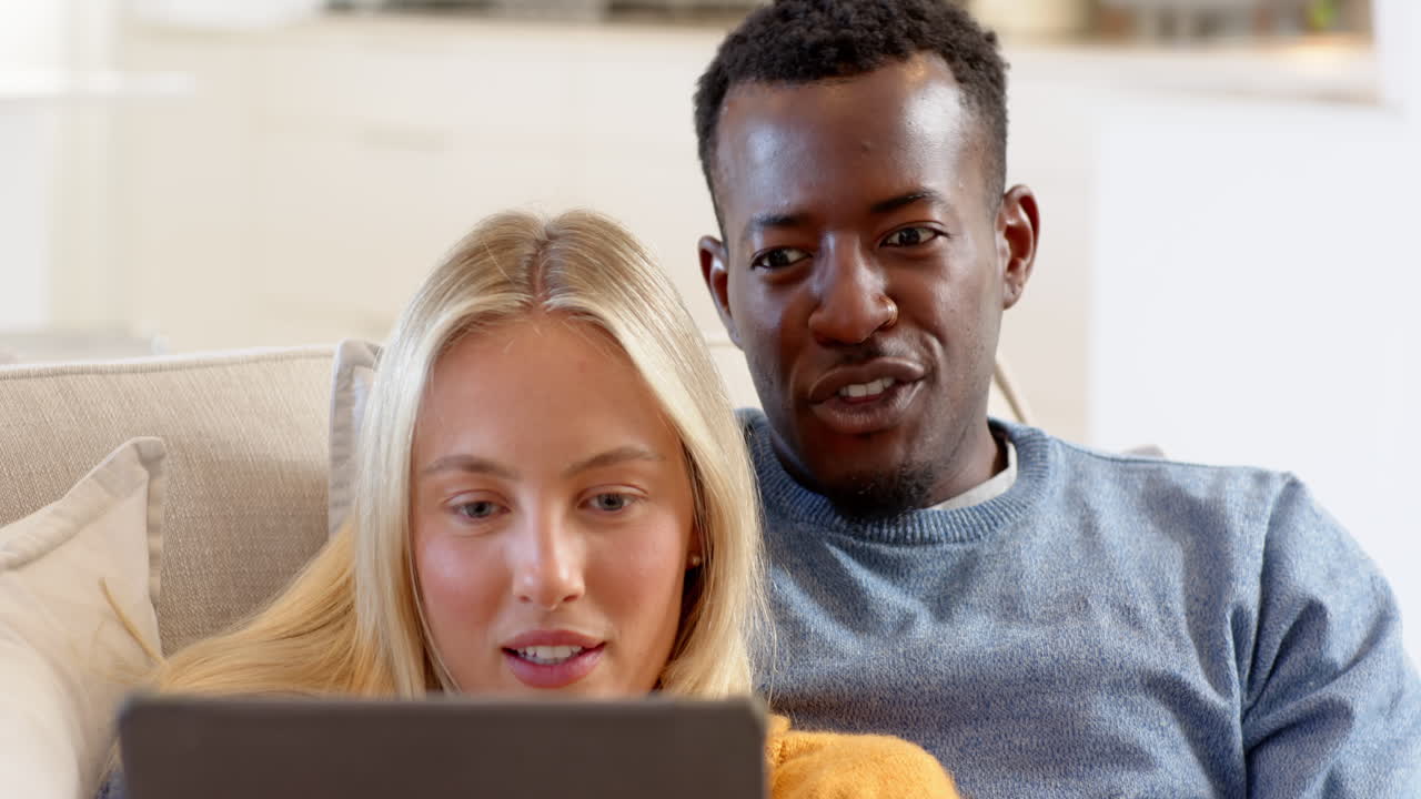 diverse couple relaxing on couch watching tablet, enjoying cozy holiday time together