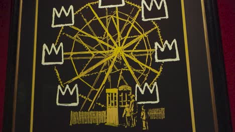 golden ferris wheel with crowns as seats at banksy exhibition in estonia tallinn. the painting or poster is well lit under lights and on a red wall. painting is framed in a black frame indoors.