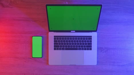 top down view of a laptop computer with mock up green screen chromakey display on a wooden office desk next to notebook with pens, glasses, and a glass of water. slow zoom out, close up