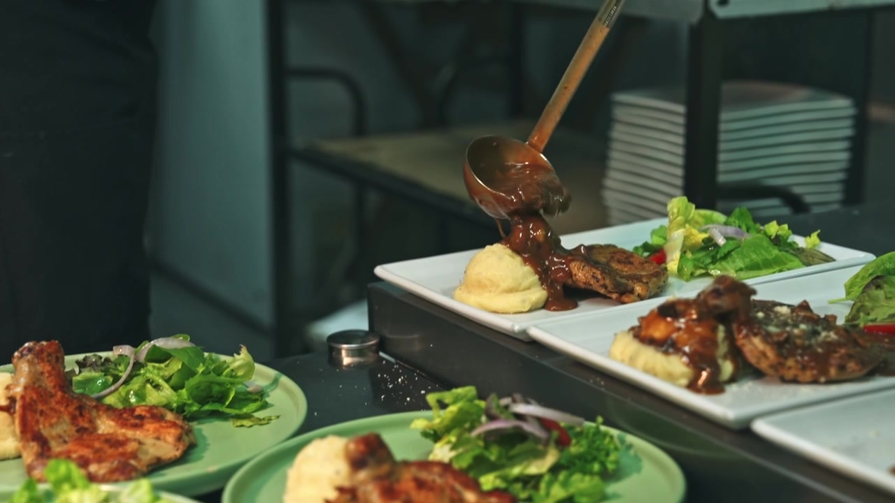 Medium tracking shot of a kitchen team garnishing, ladling sauce, and picking up finished rectangular chicken chop plates from a stainless steel pass.