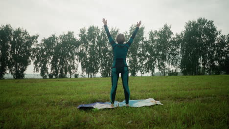 vista trasera de una mujer de pie con la cabeza ligeramente levantada, levantando las manos lentamente extendidas, luego bajándolas cerca de su cara mientras practica yoga en un vasto campo abierto bajo un cielo nublado