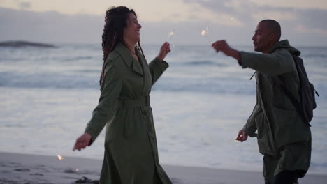 happy-couple-on-beach-dancing-celebrating-new-years-eve-waving-sparklers-enjoying-silly-fun-together-in-calm-ocean-seaside-background-at-sunset