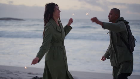 happy couple on beach dancing celebrating new years eve waving sparklers enjoying silly fun together in calm ocean seaside background at sunset