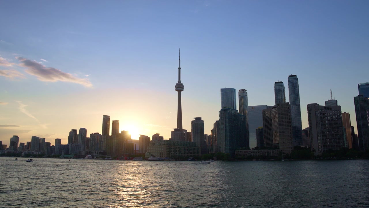 Ampia vista dello skyline di Toronto e della CN Tower da una barca in movimento mentre il sole tramonta tra gli edifici con riflessi sull'acqua, in slow motion.