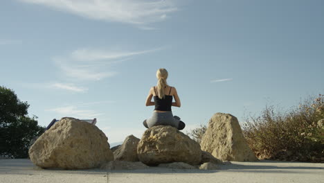 slow motion video of young blonde woman meditating on rock outside in yoga pose with blue sky