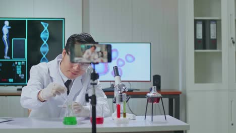 asian scientist man taking solution from test tube with micropipette putting in petri dish and using smartphone on tripod recording video