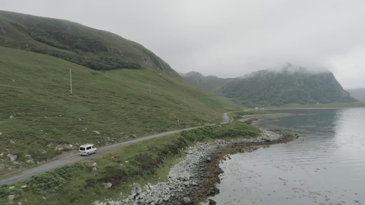 Drone tracking shot of a campervan on a single track road during a trip of the Scotland North Coast 500 with a Loch and cloudy topped mountains in view
