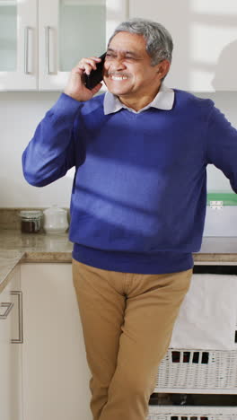 video vertical de un hombre bi-racial senior feliz hablando en su teléfono inteligente de pie en la cocina, sonriendo