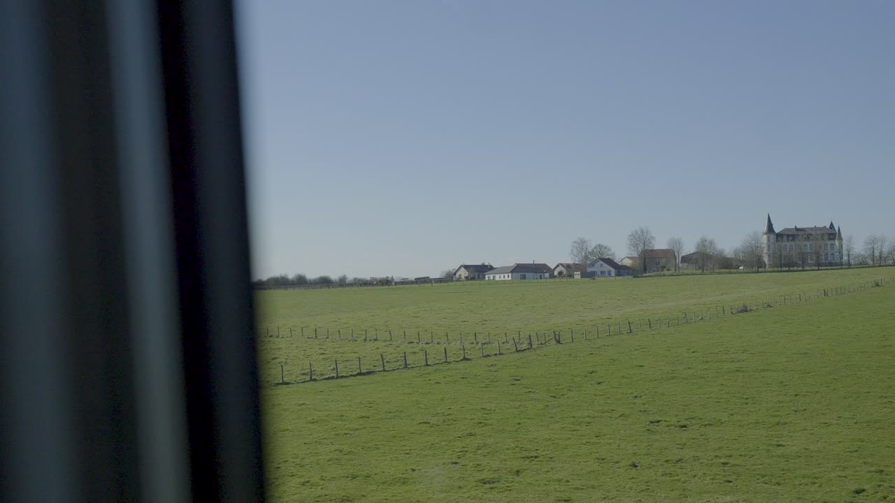 Train moving through open fields, green landscape with distant houses visible