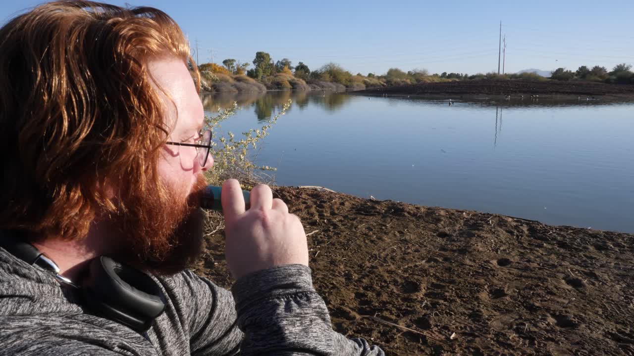 Red hair man vaping at the edge of a pond.
