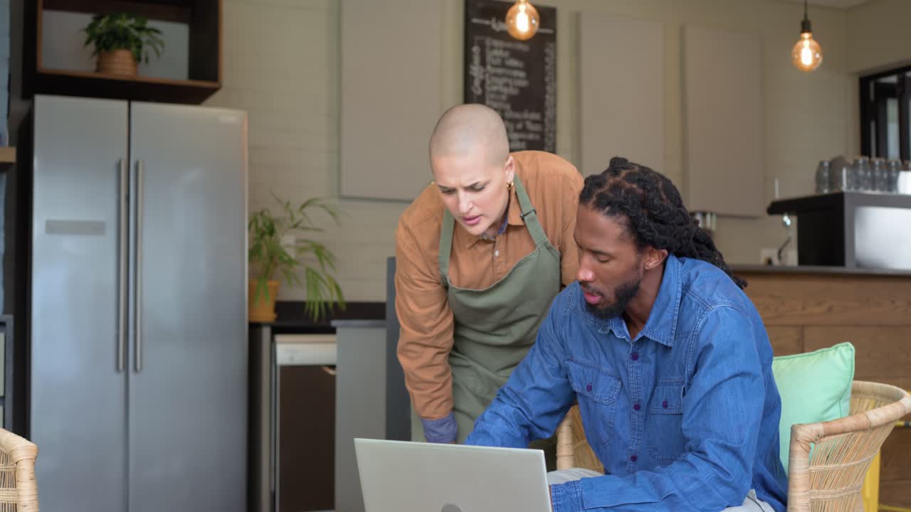 African American man typing laptop waving White woman barista approaching explaining screen in cafe