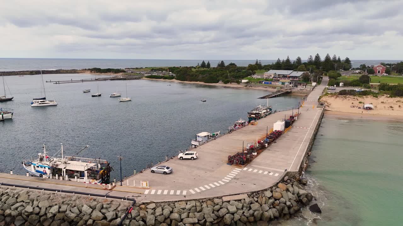 Drone footage captures Apollo Bay's serene harbor, showcasing boats, a jetty, and a rocky shoreline under overcast skies