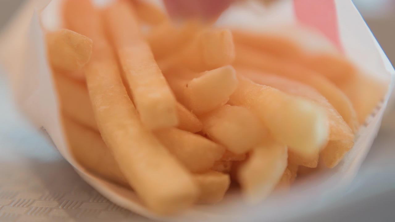 Low-angle close-up showing hand fingers reaching into white paper container crowded with golden crispy fries; visible finger motion creates gentle blur and slight displacement while warm light