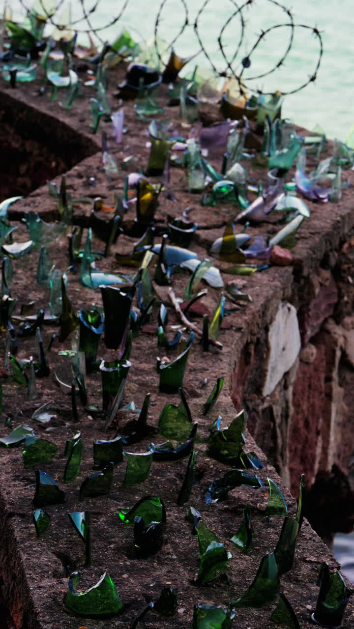 Close up of multiple broken glass shards and barbed wire loops along the edge of a brick wall with a blurred view of the sea. Vertical