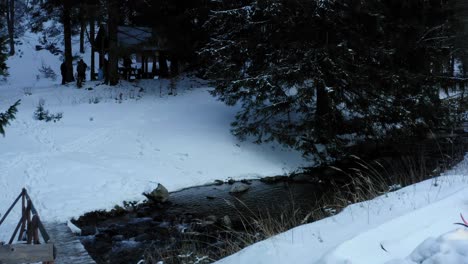 Drone-shot-over-narrow-Ladecka-river-passing-through-Bielice-mountain-range-with-the-view-of-wooden-bridge-over-the-flowing-water-in-winter-season