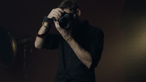 vista frontal de un joven fotógrafo caucásico tomando una foto con una cámara y sonriendo en el fondo oscuro del estudio