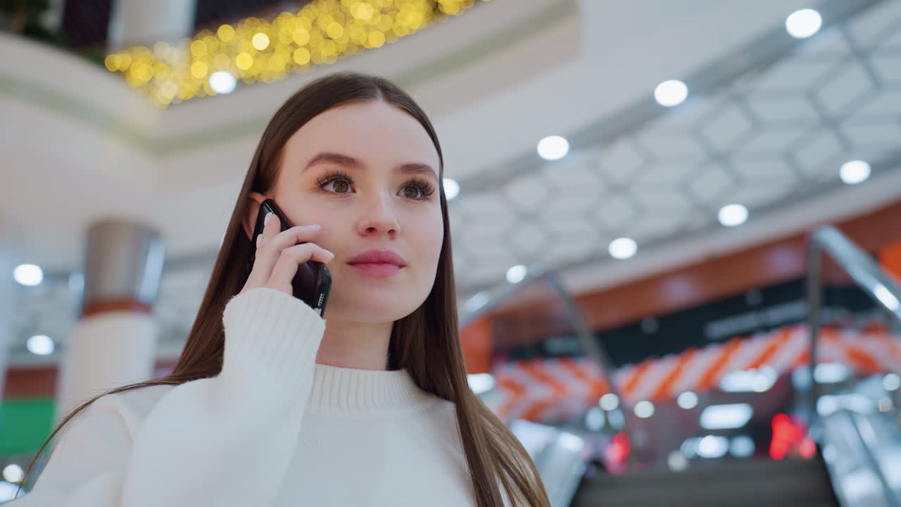 Confident young woman talking on phone while ascending escalator in stylish shopping mall, bright interior, elegant design, and warm lighting create a modern