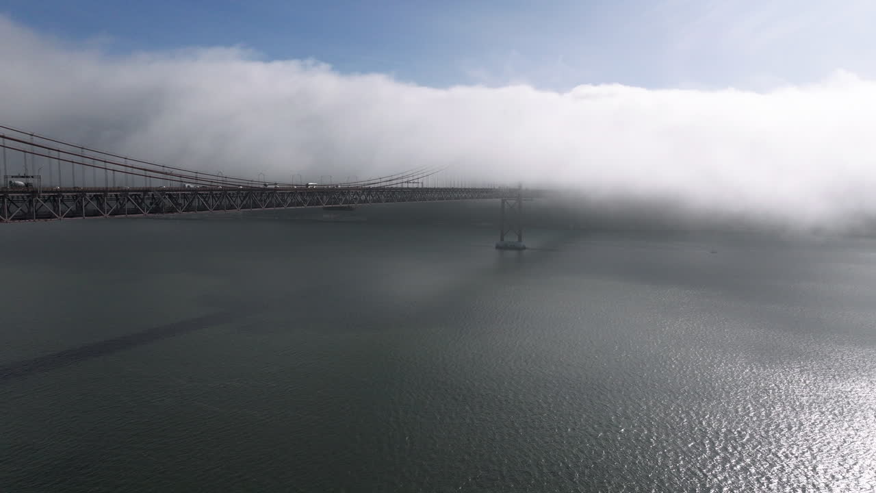 Cinematic aerial drone establishing slow shot towards the iconic red 25th April Bridge in Lisbon, Portugal, Europe. Sunny and bright with clouds, fog, mist, covering the bridge
