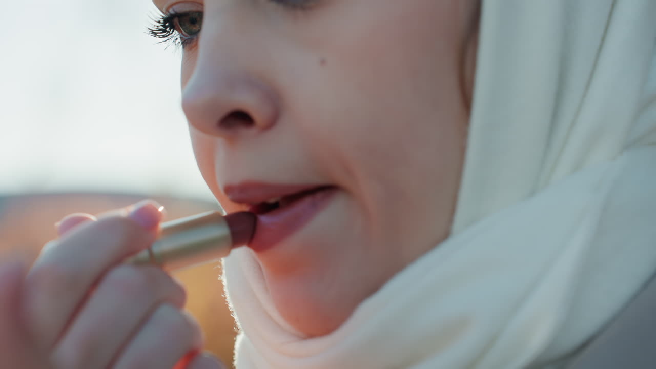 Caucasian woman closeup applying lipstick in soft autumn light, white scarf wrapped, city skyline blurred, reflective expression, subtle glitter eye makeup, careful lip application, morning ritual