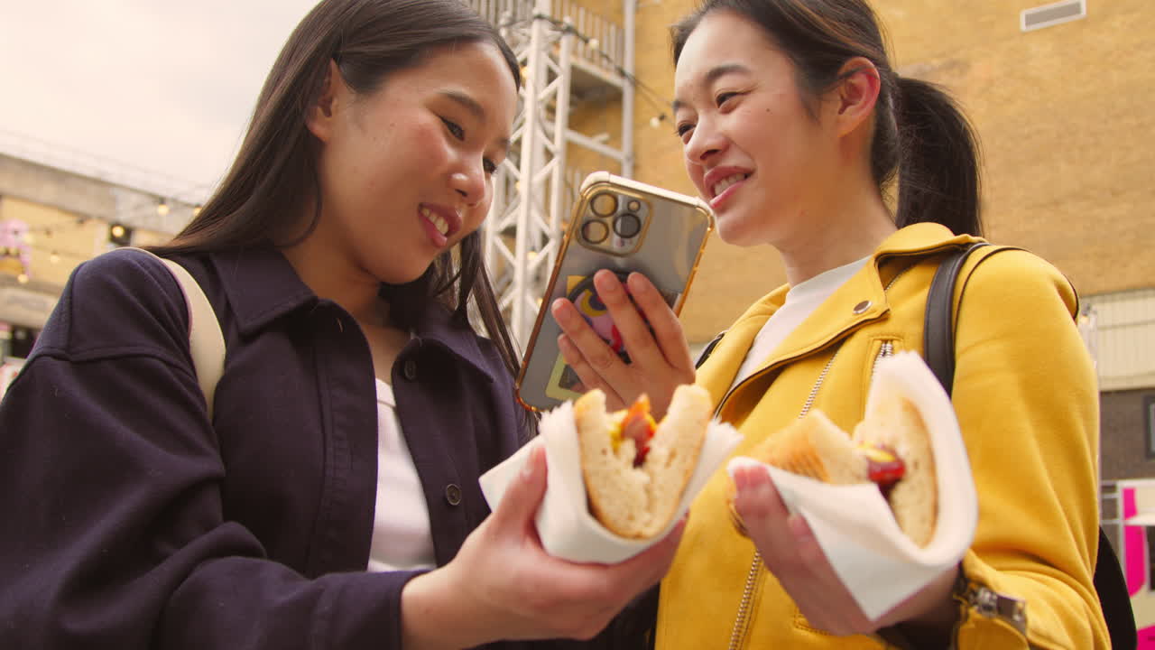 두 명의 여성 친구가 휴대 전화 2에서 거리 음식 상점에서 구입한 핫도그의 사진을 찍고 있습니다.