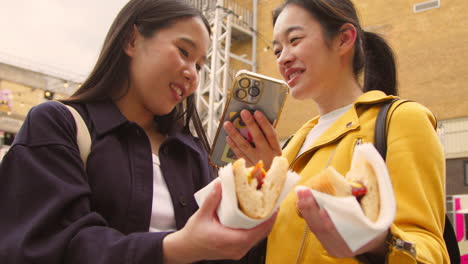 two female friends taking photo of hot dogs bought at street food stall on mobile phone 2