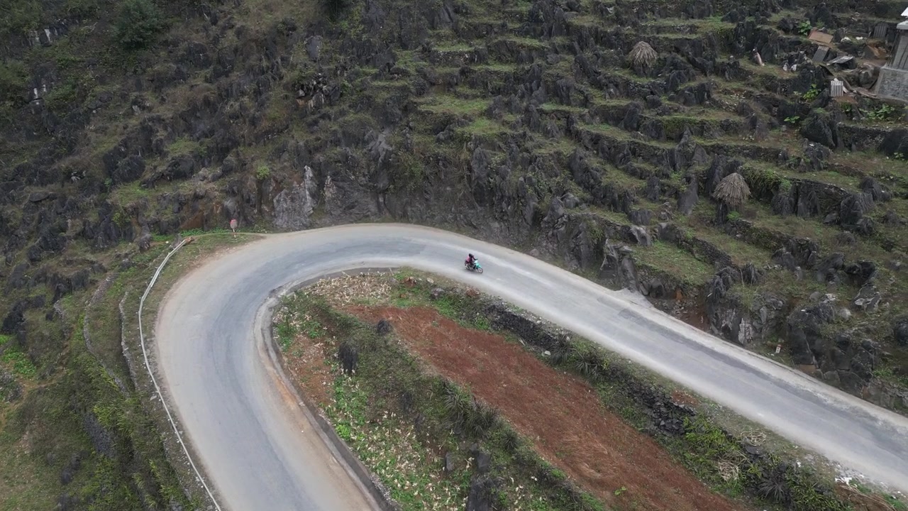 Vista aerea dall'alto con drone di un motociclista che percorre una tortuosa strada di montagna a tornanti attraverso campi terrazzati e un aspro terreno calcareo al Passo Ma Pi Leng ad Ha Giang, Vietnam