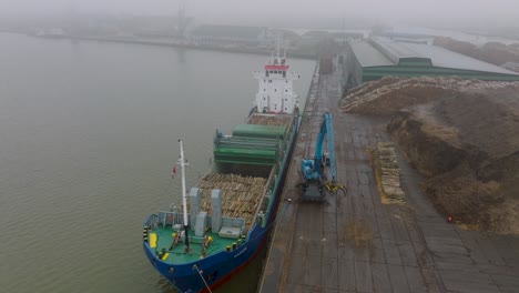 Aerial-establishing-view-of-wood-terminal-crane-loading-timber-into-the-cargo-ship,-Port-of-Liepaja-,-lumber-log-export,-overcast-day-with-fog-and-mist,-drone-shot-moving-forward,-tilt-down