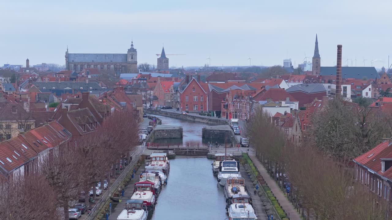 Úchvatný vzlet dronu začínající nízko nad historickým kanálem v Bruggách, stoupající, aby odhalil ikonické vlámské střechy, kostelní věže a rozsáhlé městské panorama zapsané na seznamu světového dědictví UNESCO.