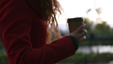 young woman in red coat is drinking coffee on the street while walking on cold autumn day